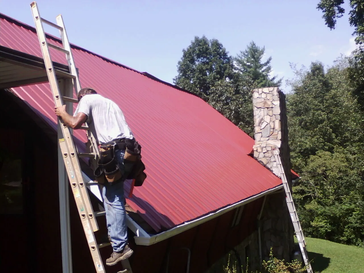Roofer on a ladder beside a red metal roof and stone chimney — Soffit & Fascia Repair by Roofing Brothers in Middle River